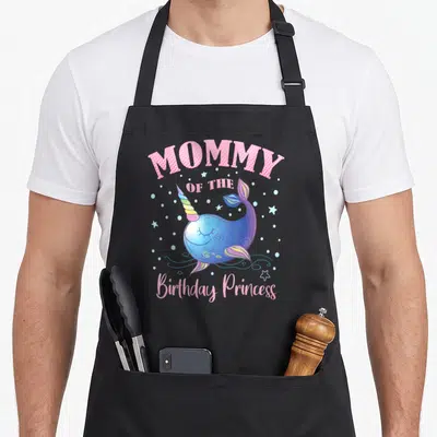 Narwhal princess apron - MOMMY OF THE Birthday Princess text surrounding a blue cartoon narwhal with rainbow horn — shown on a black cotton canvas apron against a white background