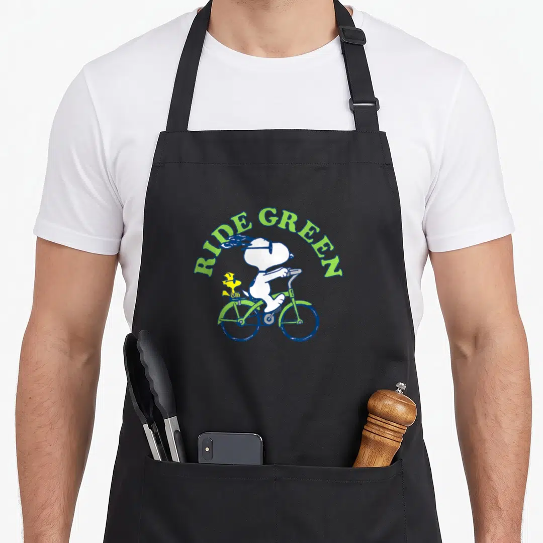 Funny ride novelty apron - Snoopy riding a green bicycle with Woodstock under arched RIDE GREEN text in cartoon style — shown on a black cotton canvas apron against a white background