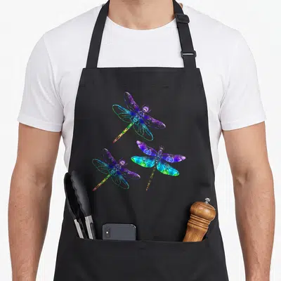 Wrinkled hippie apron - Three rainbow dragonflies in purple blue green yellow red cartoon style — shown on a black cotton canvas apron against a white background