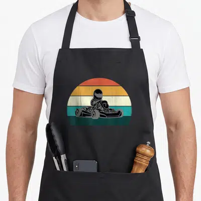 Motor karting apron - Retro go-kart driver silhouette on striped semi-circle background in red orange teal and green — shown on a black cotton canvas apron against a white background