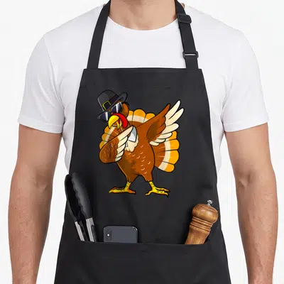 Dabbing turkey pilgrim thanksgiving day aprons - Cartoon brown turkey wearing black pilgrim hat and sunglasses with one — shown on a black cotton canvas apron against a white background