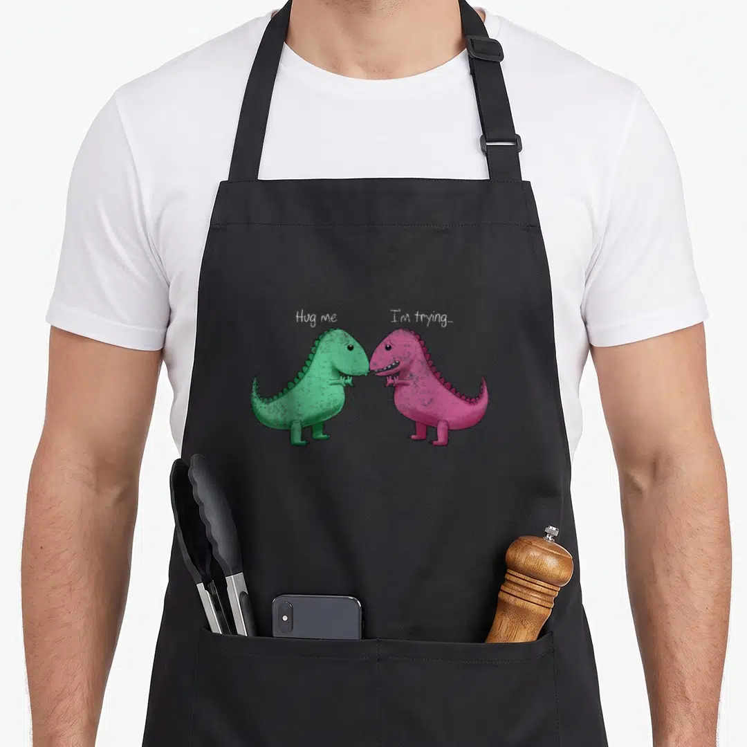 Hug dinosaur apron - Two facing cartoon dinosaurs in green and pink with text 'Hug me, I'm trying...' — shown on a black cotton canvas apron against a white background