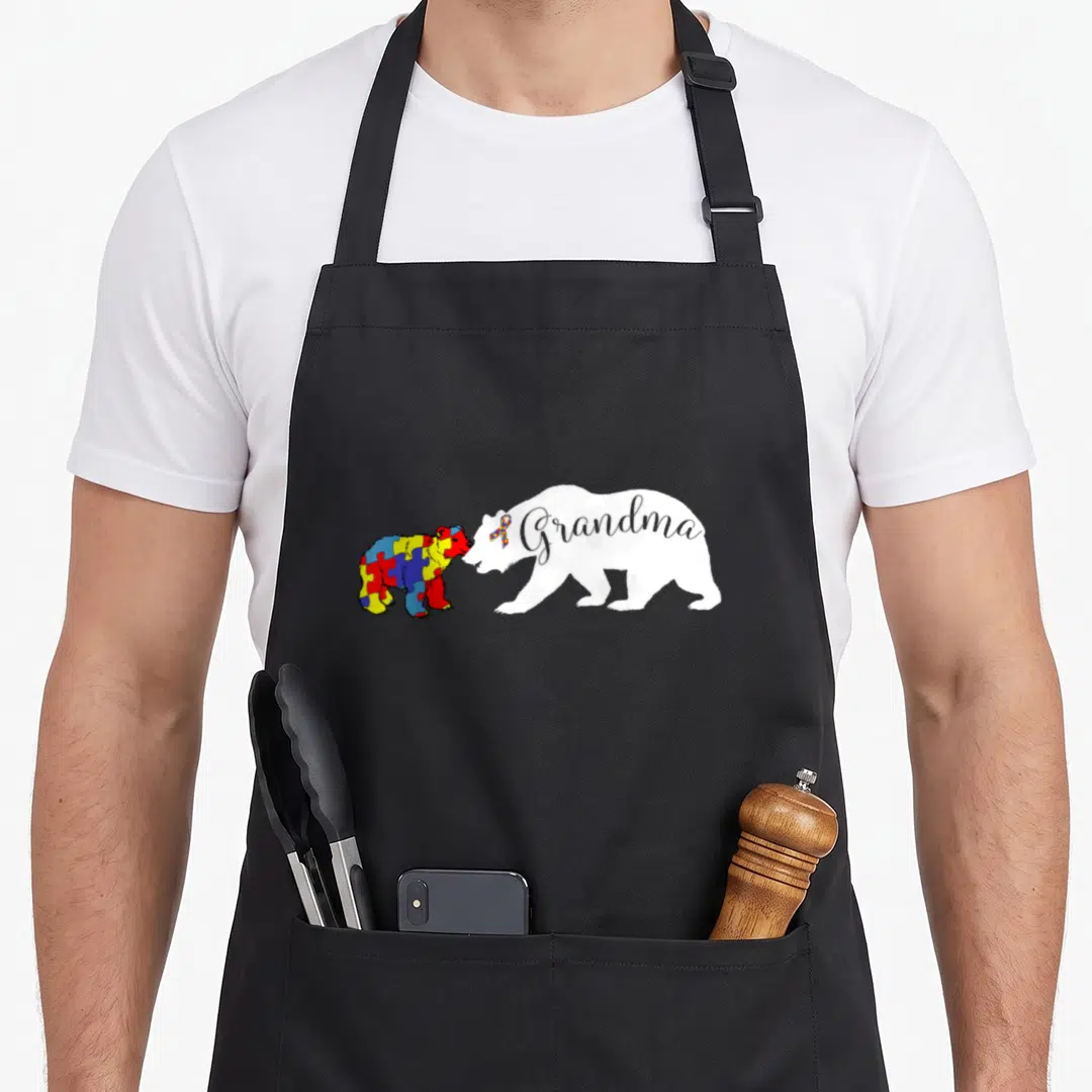 Grandma cute bear apron - White bear silhouette with Grandma text nuzzling a blue yellow red grey puzzle pattern cub — shown on a black cotton canvas apron against a white background