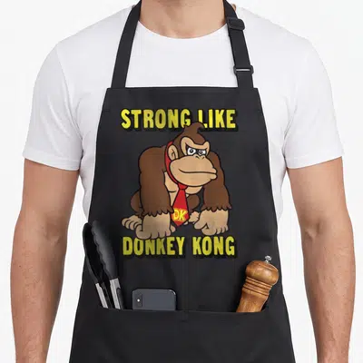 Strong donkey apron - Brown gorilla wearing red tie with DK initials between yellow text lines — shown on a black cotton canvas apron against a white background