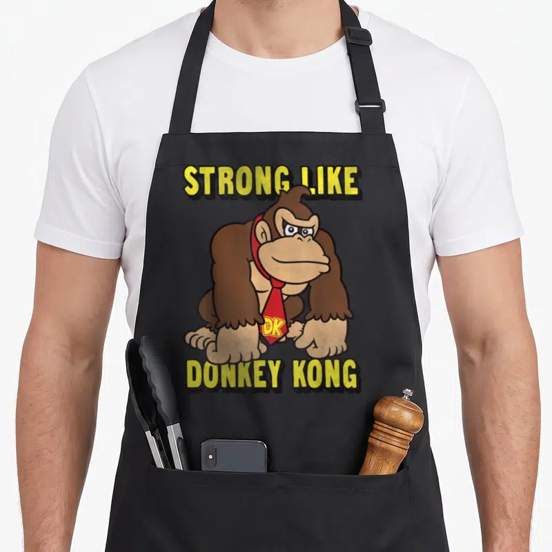 Strong donkey apron - Brown gorilla wearing red tie with DK initials between yellow text lines — shown on a black cotton canvas apron against a white background
