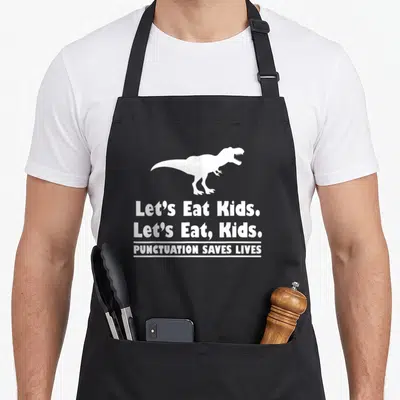 Dinosaur let's eat apron - White T-Rex silhouette above stacked text Let's Eat Kids and Punctuation Saves Lives — shown on a black cotton canvas apron against a white background