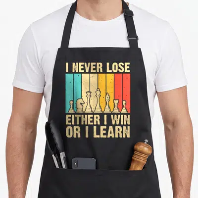 Cute chess player apron - Retro striped graphic with chess pieces and text I Never Lose Either I Win Or I Learn — shown on a black cotton canvas apron against a white background