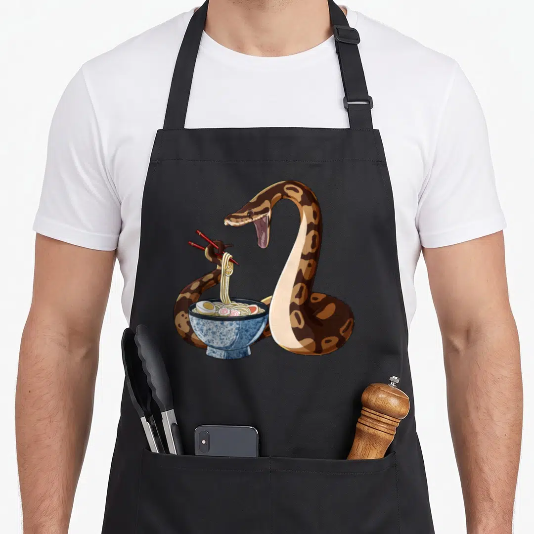 Snake anime apron - Cartoon brown python holding red chopsticks lifting noodles from a blue bowl — shown on a black cotton canvas apron against a white background