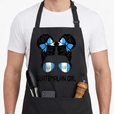 Guatemalan messy aprons - Back of girl head with two buns, flag sunglasses, and GUATEMALAN GIRL text — shown on a black cotton canvas apron against a white background