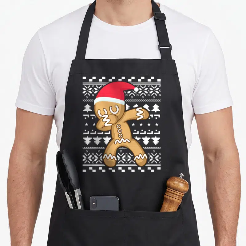 Dabbing gingerbread apron - Cartoon gingerbread man dabbing in red Santa hat on white snowflake fair-isle pattern — shown on a black cotton canvas apron against a white background