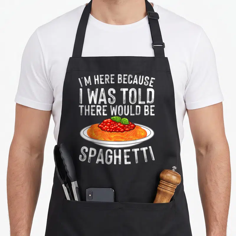 Cute spaghetti pasta apron - Cartoon plate of orange spaghetti with red sauce and green basil under white stacked text — shown on a black cotton canvas apron against a white background