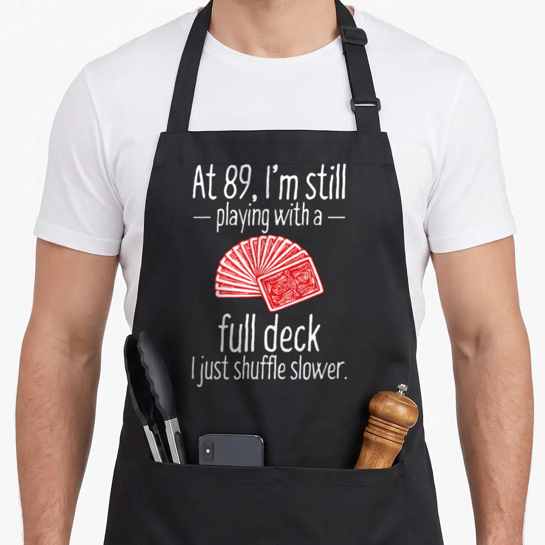 89th birthday apron - White text reading At 89 full deck shuffle slower above red fanned playing cards — shown on a black cotton canvas apron against a white background
