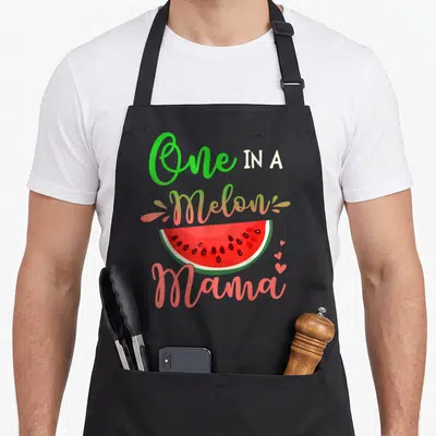 Melon mama apron - One IN A Melon Mama text with watermelon slice and three hearts — shown on a black cotton canvas apron against a white background