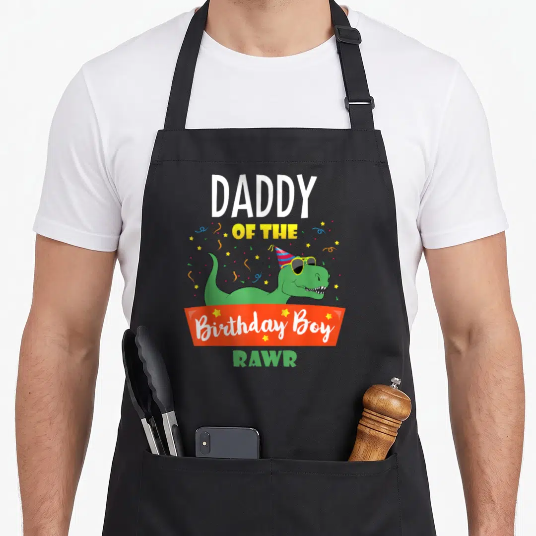 Daddy family cooking aprons - Green dinosaur in sunglasses and party hat with Daddy of the Birthday Boy text — shown on a black cotton canvas apron against a white background