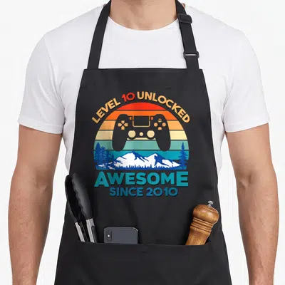 Level unlocked birthday apron - Retro game controller over striped sun with mountain range and pine trees — shown on a black cotton canvas apron against a white background