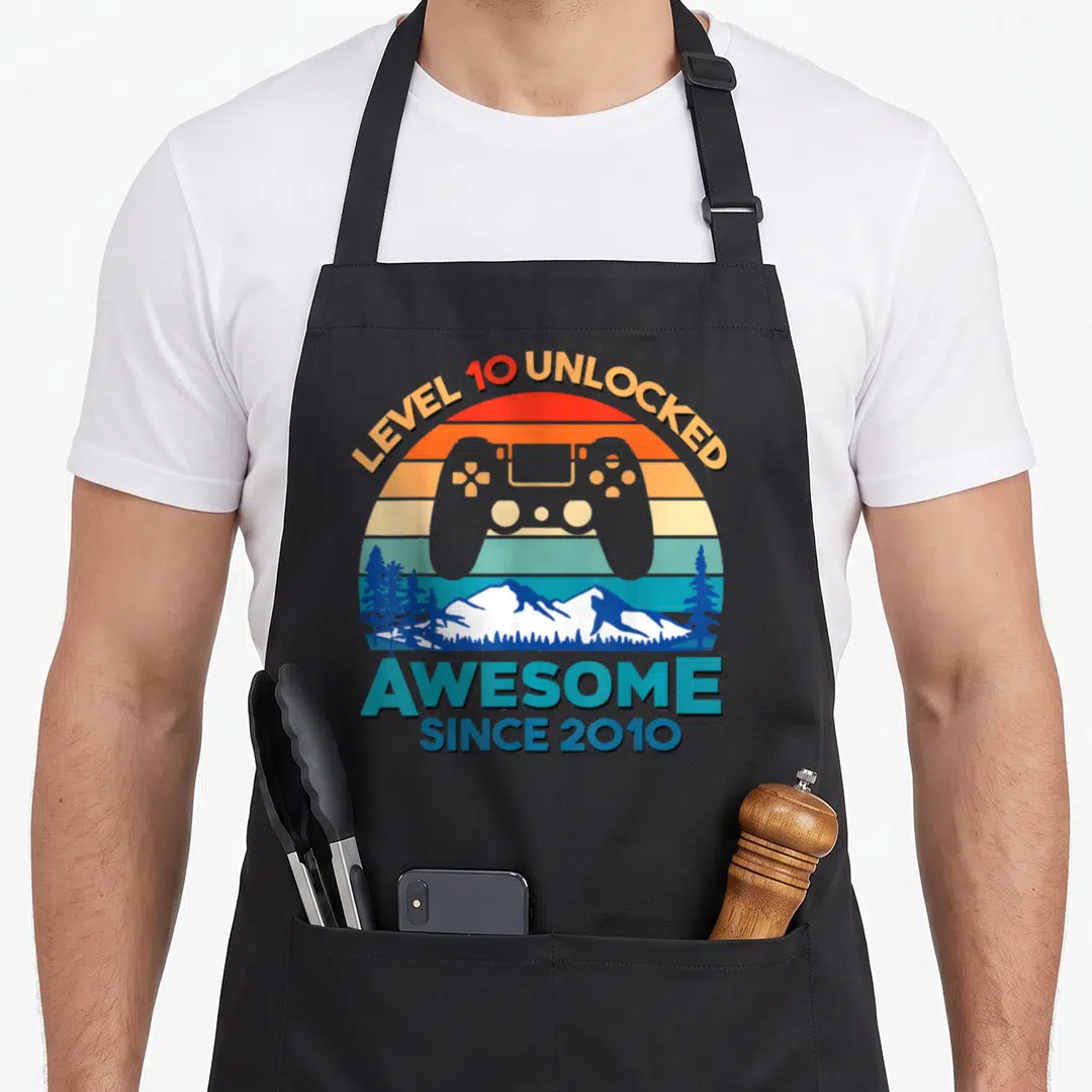 Level unlocked birthday apron - Retro game controller over striped sun with mountain range and pine trees — shown on a black cotton canvas apron against a white background