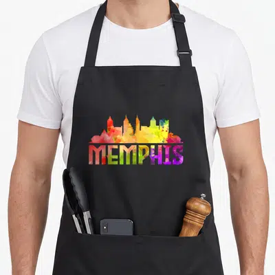 Skyline memphis apron - Multicolored watercolor Memphis skyline silhouette above matching text in red, yellow, green — shown on a black cotton canvas apron against a white background