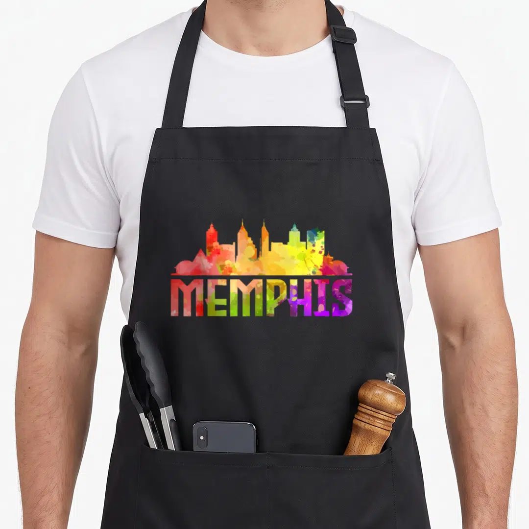 Skyline memphis apron - Multicolored watercolor Memphis skyline silhouette above matching text in red, yellow, green — shown on a black cotton canvas apron against a white background