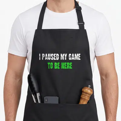 Video game gaming apron - Distressed white and green text reading I PAUSED MY GAME TO BE HERE on black fabric — shown on a black cotton canvas apron against a white background