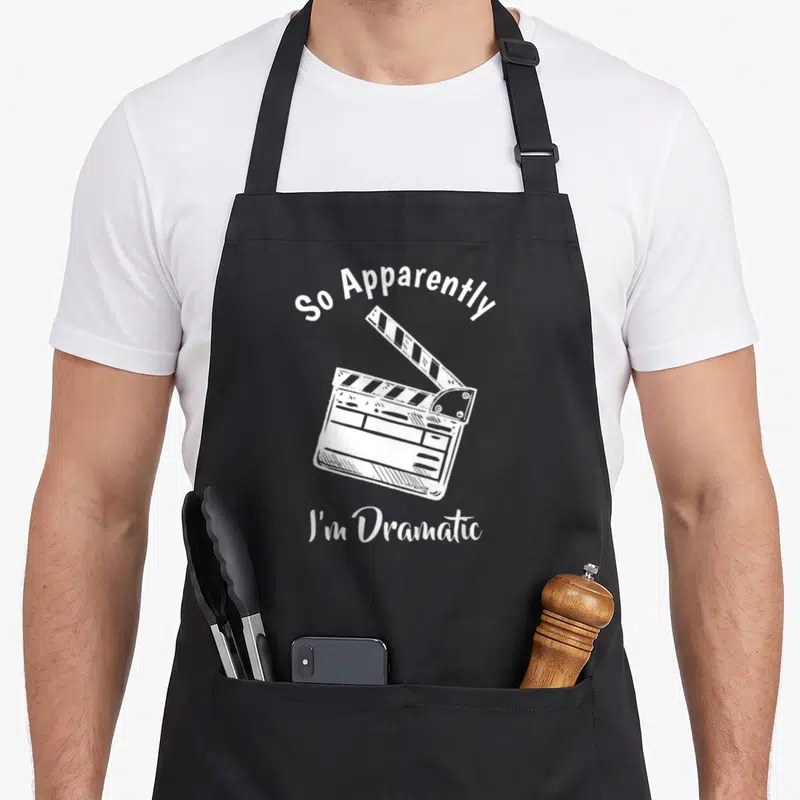 Apparently acting apron - White clapperboard illustration with text So Apparently I'm Dramatic — shown on a black cotton canvas apron against a white background