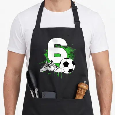 6th soccer birthday apron - Number 6 with soccer ball and cleats on green splatter — shown on a black cotton canvas apron against a white background