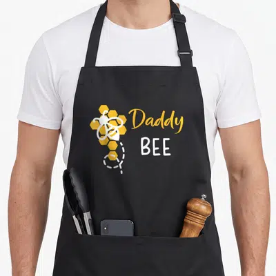 1st bee aprons - Daddy BEE text with cartoon bee and honeycomb in yellow and white — shown on a black cotton canvas apron against a white background