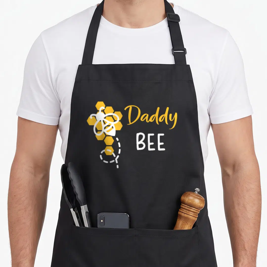 1st bee aprons - Daddy BEE text with cartoon bee and honeycomb in yellow and white — shown on a black cotton canvas apron against a white background