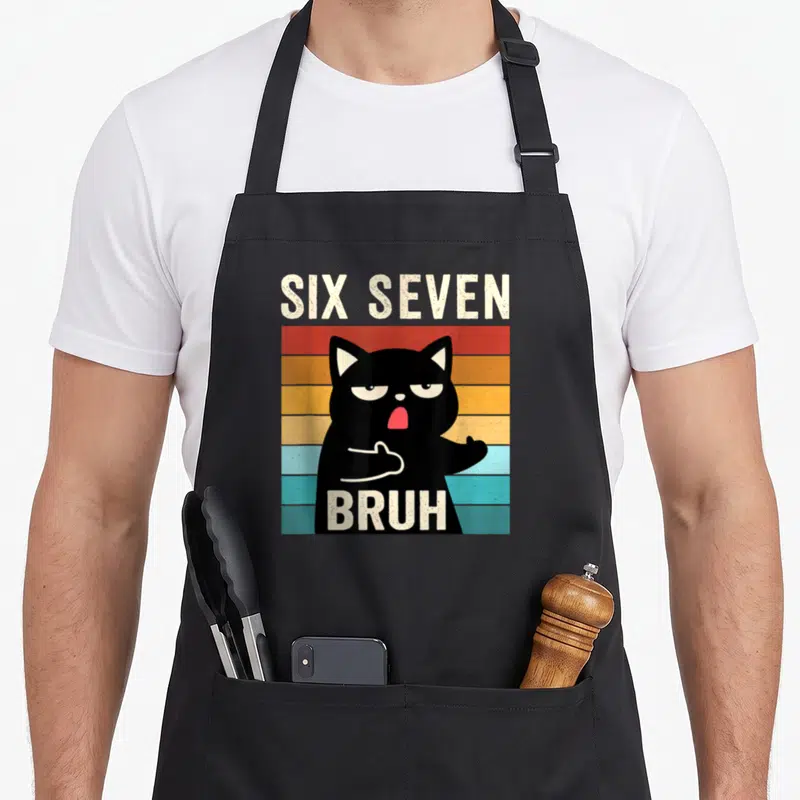 Seven cat apron - Black cat over multi-colored stripes with six seven bruh text in retro style — shown on a black cotton canvas apron against a white background