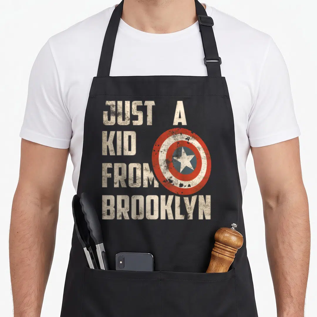Shield brooklyn apron - Stacked text JUST A KID FROM BROOKLYN beside a distressed red white and blue shield — shown on a black cotton canvas apron against a white background