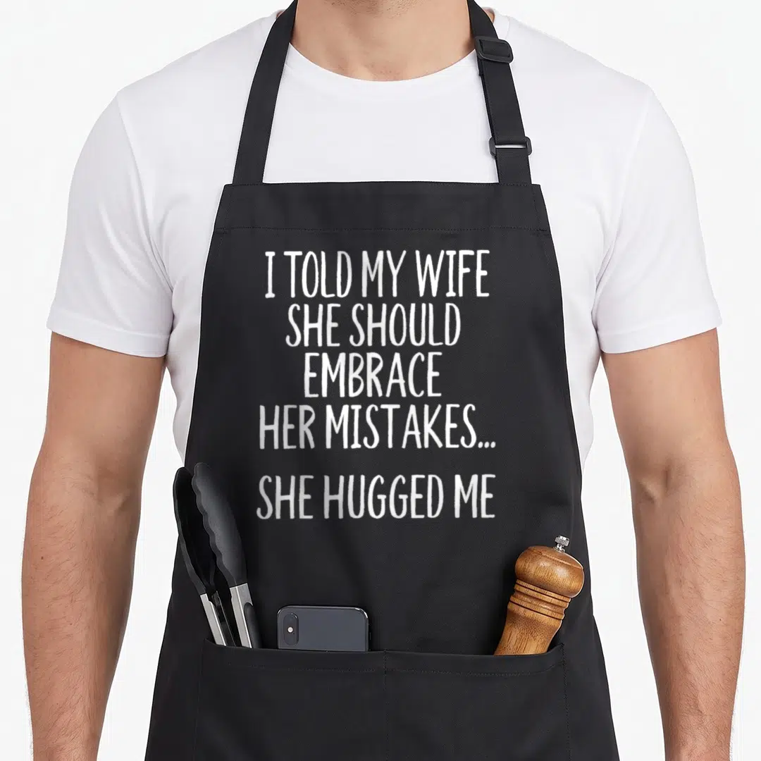 Embrace wife apron - White stacked text: I TOLD MY WIFE SHE SHOULD EMBRACE HER MISTAKES...SHE HUGGED ME — shown on a black cotton canvas apron against a white background