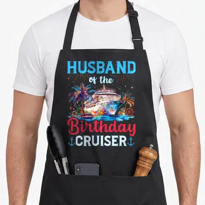 Cruise husband apron - Cruise ship graphic with HUSBAND of the Birthday CRUISER text, palm trees, and anchors — shown on a black cotton canvas apron against a white background