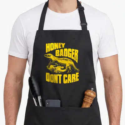 Ripple badger apron - Yellow honey badger illustration with arched text honey badger don't care in distressed font — shown on a black cotton canvas apron against a white background