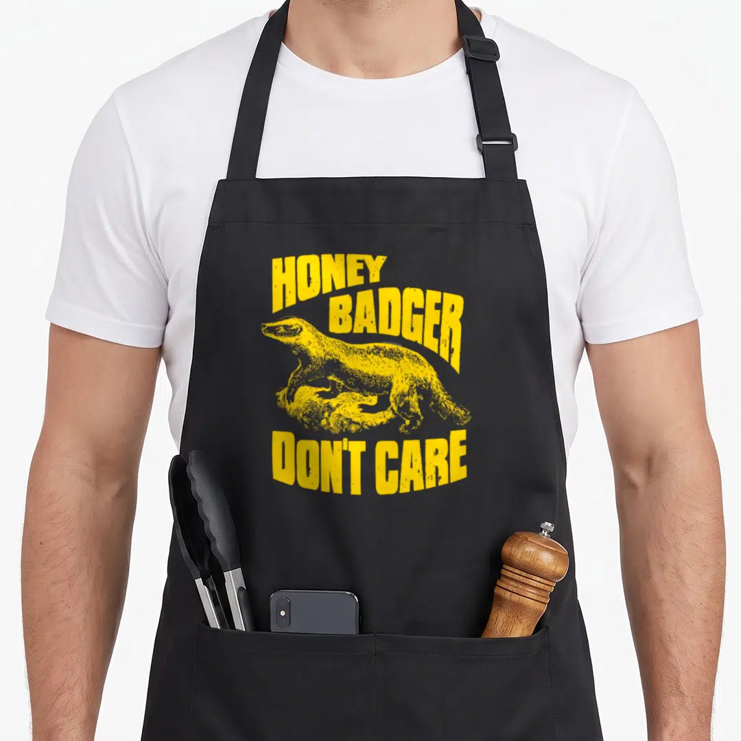 Ripple badger apron - Yellow honey badger illustration with arched text honey badger don't care in distressed font — shown on a black cotton canvas apron against a white background