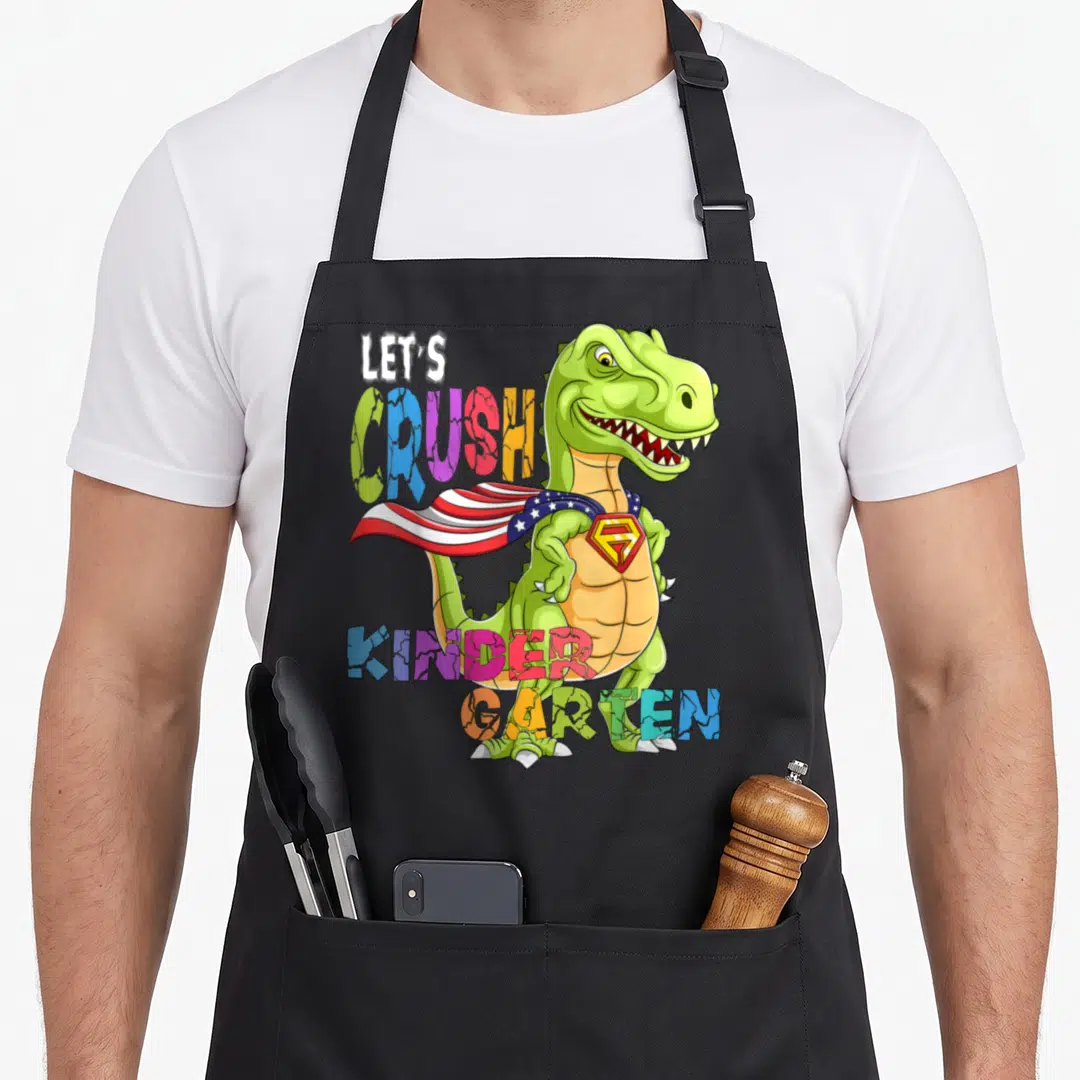Ready crush kindergarten dinosaur apron - Green T-Rex superhero with colorful LETS CRUSH KINDERGARTEN text — shown on a black cotton canvas apron against a white background