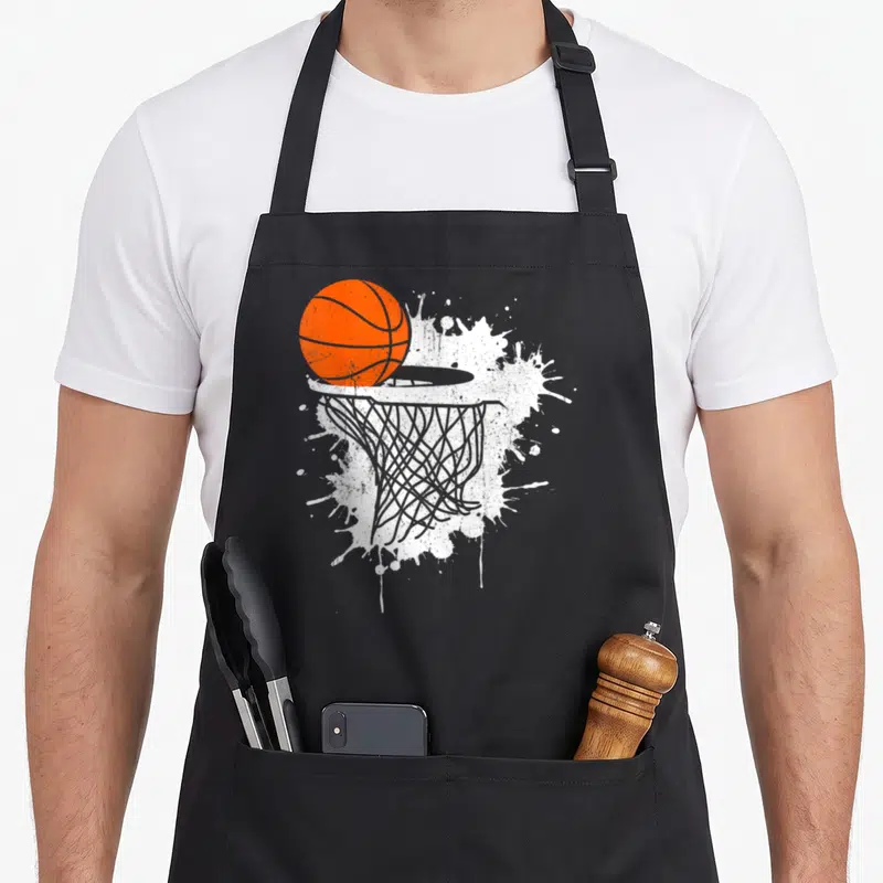 Player basketball coach apron - Orange basketball entering a hoop over a white paint splatter background in cartoon — shown on a black cotton canvas apron against a white background
