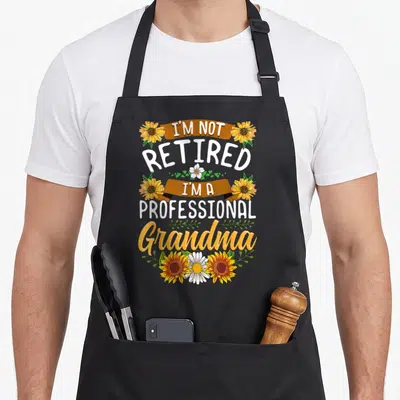 Grandma professional apron - Stacked text I'm Not Retired I'm A Professional Grandma with orange sunflowers and brown — shown on a black cotton canvas apron against a white background