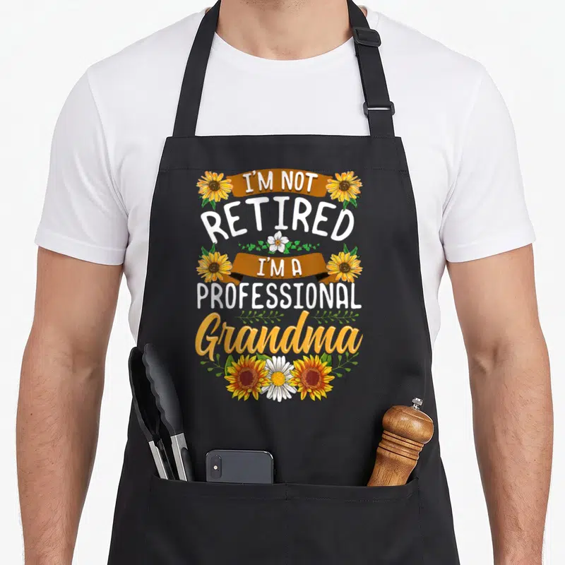 Grandma professional apron - Stacked text I'm Not Retired I'm A Professional Grandma with orange sunflowers and brown — shown on a black cotton canvas apron against a white background