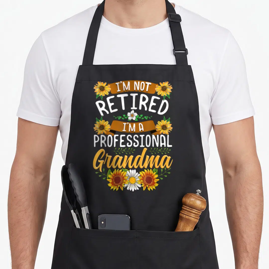 Grandma professional apron - Stacked text I'm Not Retired I'm A Professional Grandma with orange sunflowers and brown — shown on a black cotton canvas apron against a white background