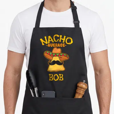 Bob taco apron - Cartoon nacho chip with sombrero and mustache under Nacho Average Bob text — shown on a black cotton canvas apron against a white background