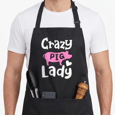 Retro crazy pig lady apron - Pink pig silhouette with PIG inside, surrounded by white Crazy and Lady text and a small — shown on a black cotton canvas apron against a white background