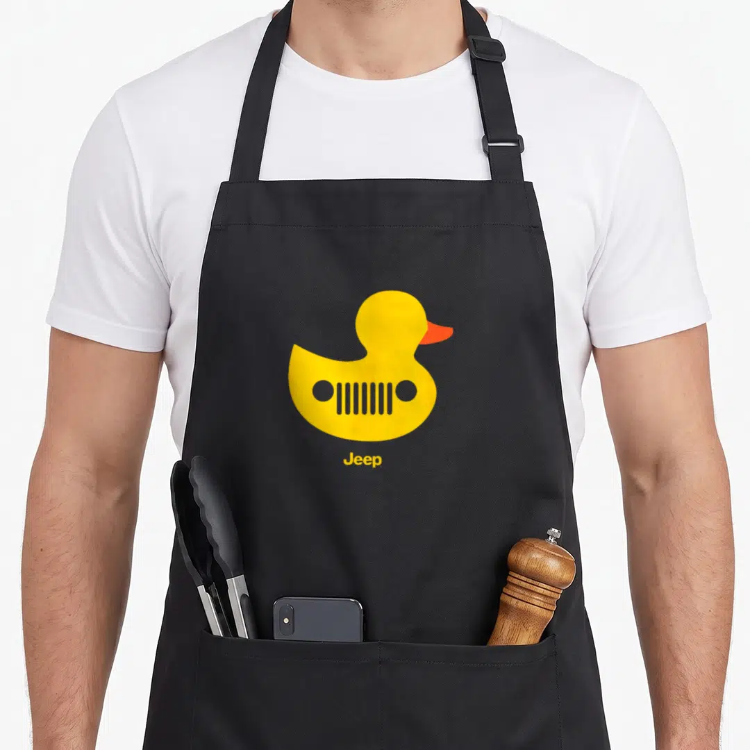 Jeep duck apron - Yellow rubber duck with grille cutouts and orange beak above text Jeep — shown on a black cotton canvas apron against a white background
