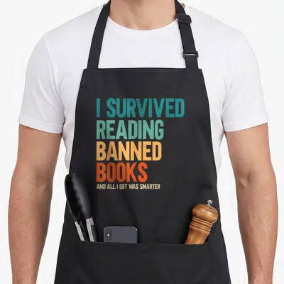 Survived reading book apron - Stacked colorful text reading I survived reading banned books and all I got was smarter — shown on a black cotton canvas apron against a white background