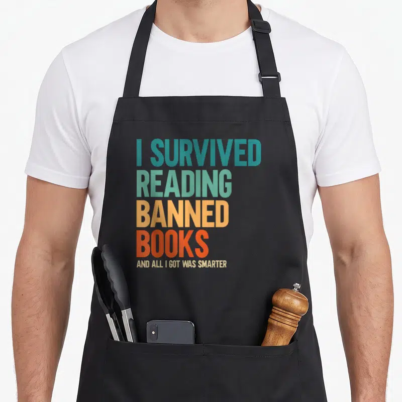 Survived reading book apron - Stacked colorful text reading I survived reading banned books and all I got was smarter — shown on a black cotton canvas apron against a white background