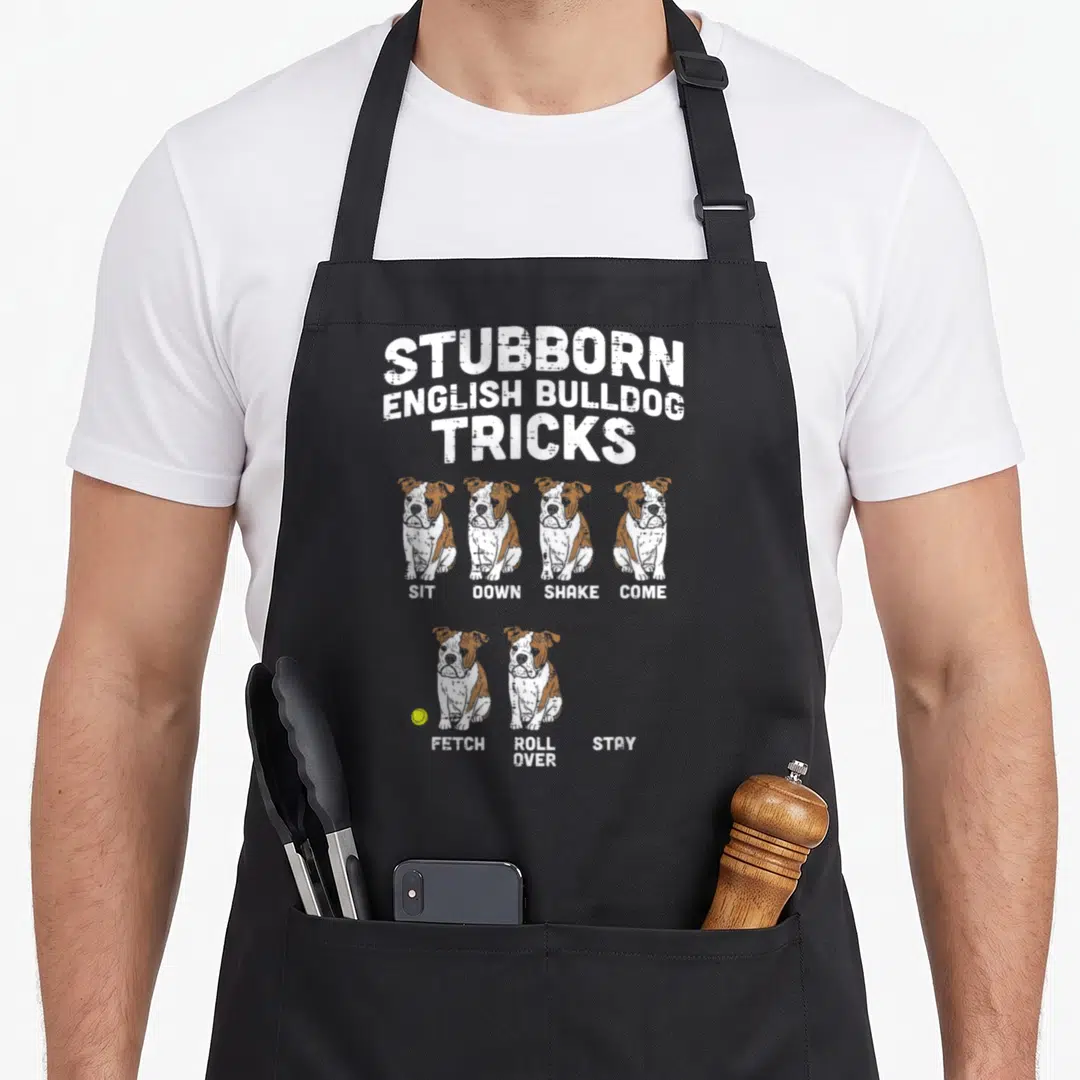 Funny tricks apron - Stubborn English Bulldog Tricks text with six sitting dogs and commands including Sit, Down — shown on a black cotton canvas apron against a white background