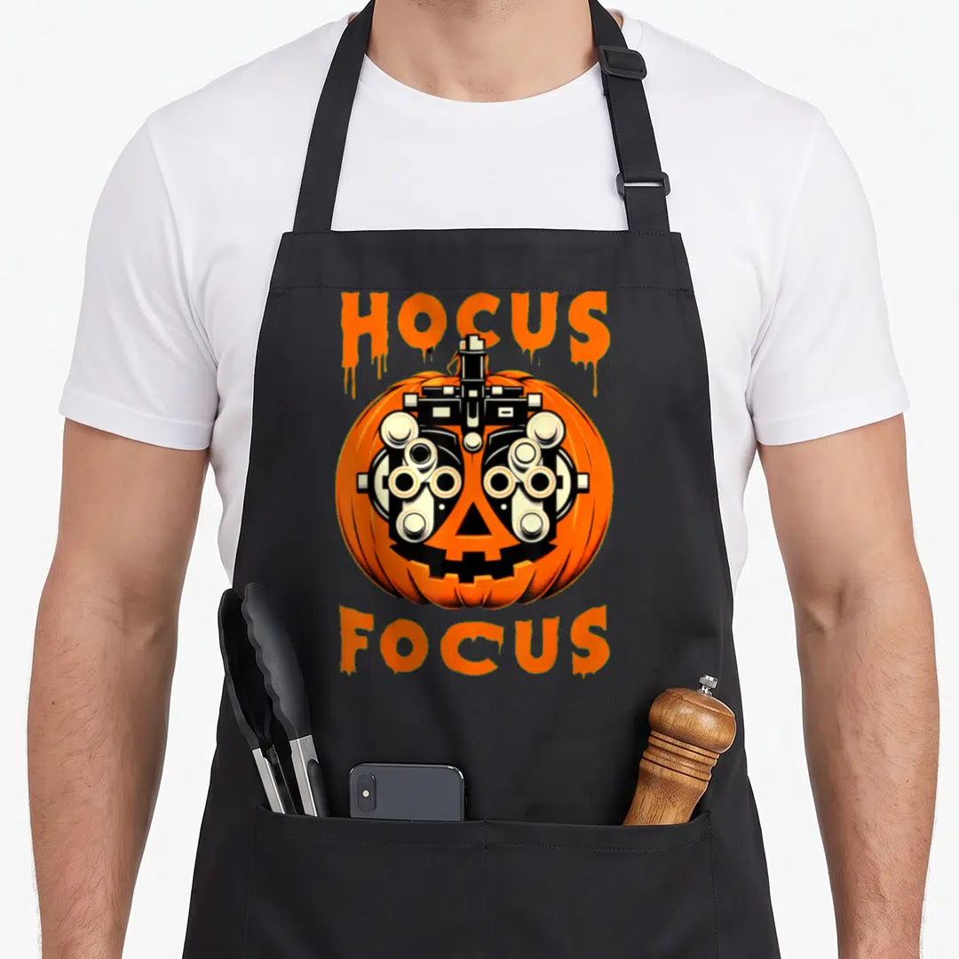 Optometry halloween apron - Orange pumpkin with phoropter eyes and Hocus Focus text in dripping font — shown on a black cotton canvas apron against a white background