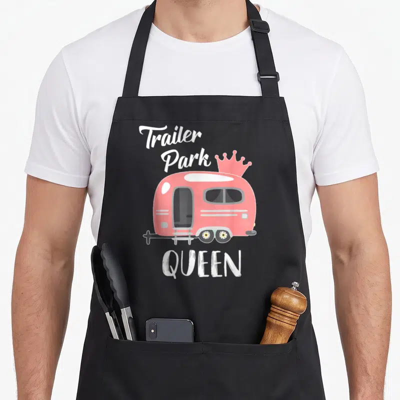 Queen trailer apron - Pink cartoon trailer with crown and white Trailer Park QUEEN text — shown on a black cotton canvas apron against a white background