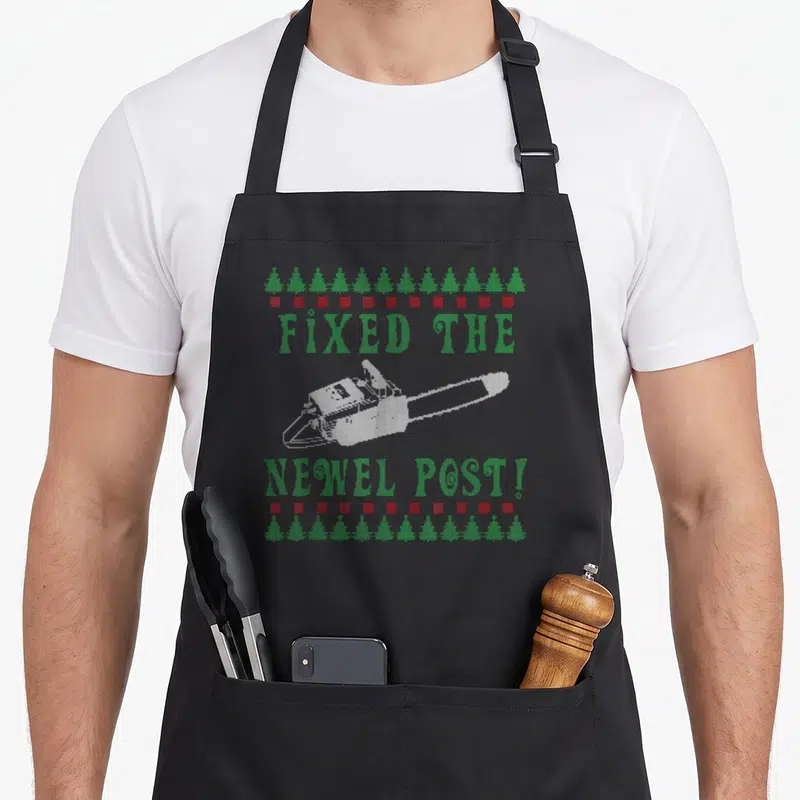 Chainsaw ugly christmas sweater apron - Pixel art chainsaw with green text Fixed the Newel Post framed by trees and red — shown on a black cotton canvas apron against a white background