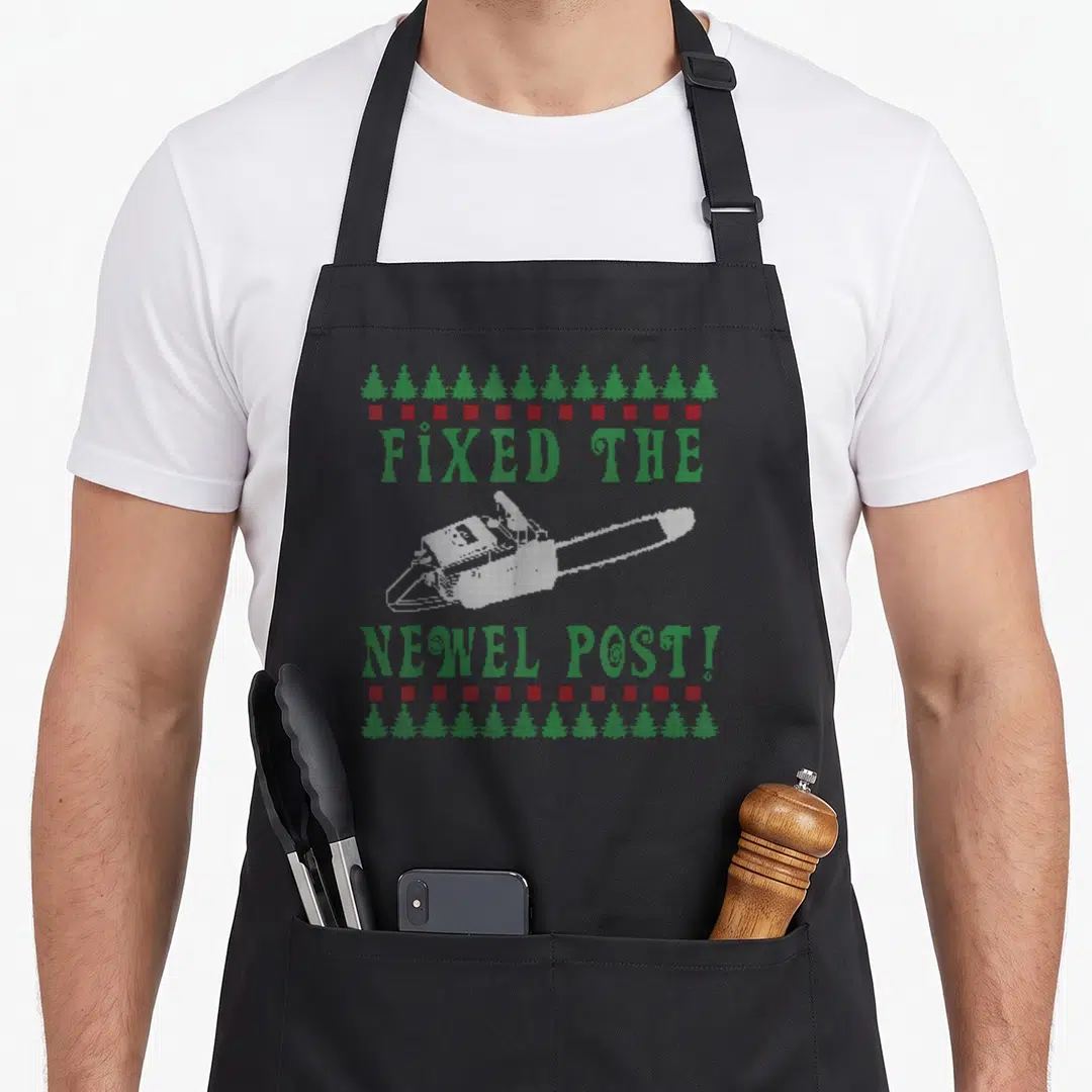 Chainsaw ugly christmas sweater apron - Pixel art chainsaw with green text Fixed the Newel Post framed by trees and red — shown on a black cotton canvas apron against a white background