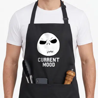 Current christmas apron - Cartoon skull with stitched mouth and stacked text reading CURRENT MOOD in white and black — shown on a black cotton canvas apron against a white background