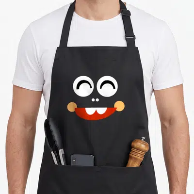 Vipkid teacher apron - Cartoon face with big eyes, orange cheeks, and red smiling mouth on grey background — shown on a black cotton canvas apron against a white background